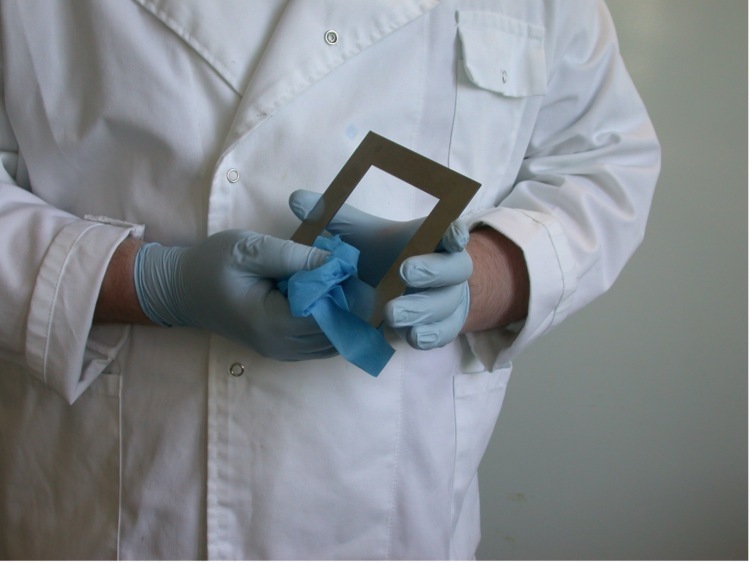 A person in a white lab coat, wiping a metal square frame called a template.