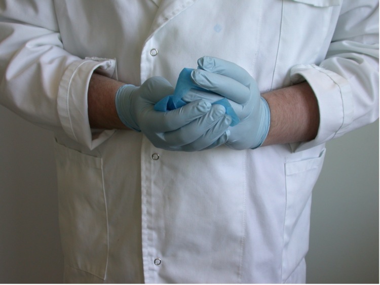 A person in a white lab coat, wearing blue latex gloves and using an alcohol wipe to clean them.