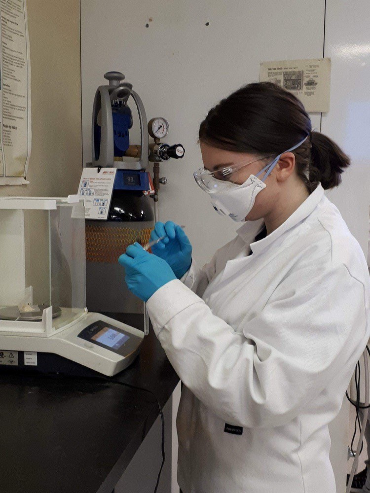 A women wearing a white laboratory jacket, wearing eye protection, a mask and blue gloves working on a sample