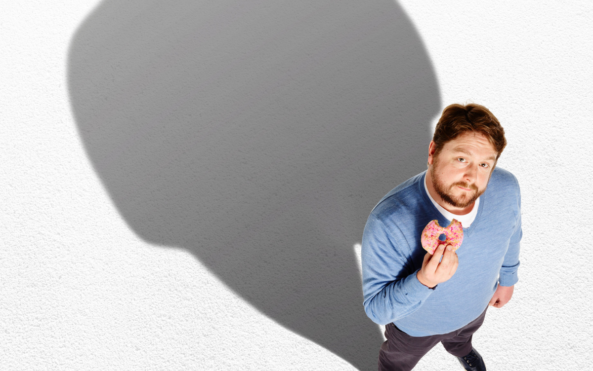 A man ion his 40s eats a a pink iced doughnut and casts a large shadow behind