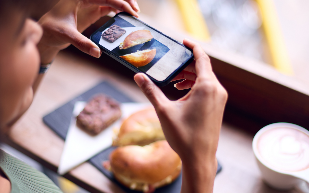 A woman is taking a photograph with the smartphone of a bagel and brownie.