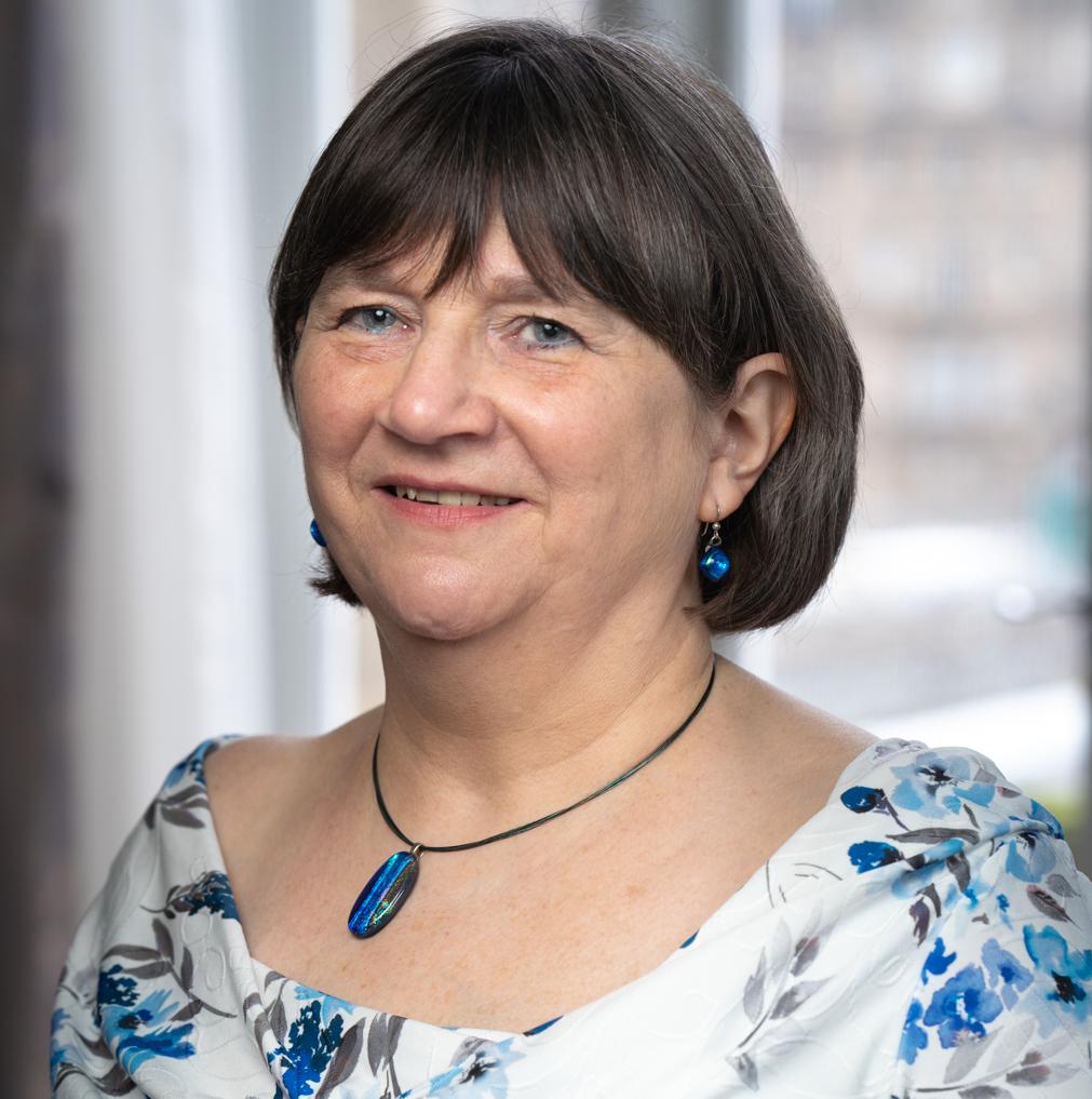 A woman with short, dark hair wearing a white blouse with blue floral patterns, a blue pendant necklace, and matching blue earrings.