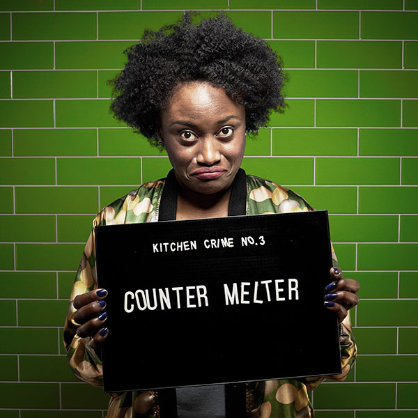 Mock mug shot photograph of a woman holding a sign saying kitchen crimes number 3: counter melter