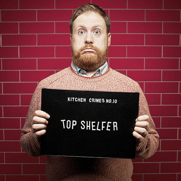 Mock mug shot photograph of a man holding a sign saying kitchen crimes number 10: top shelfer 